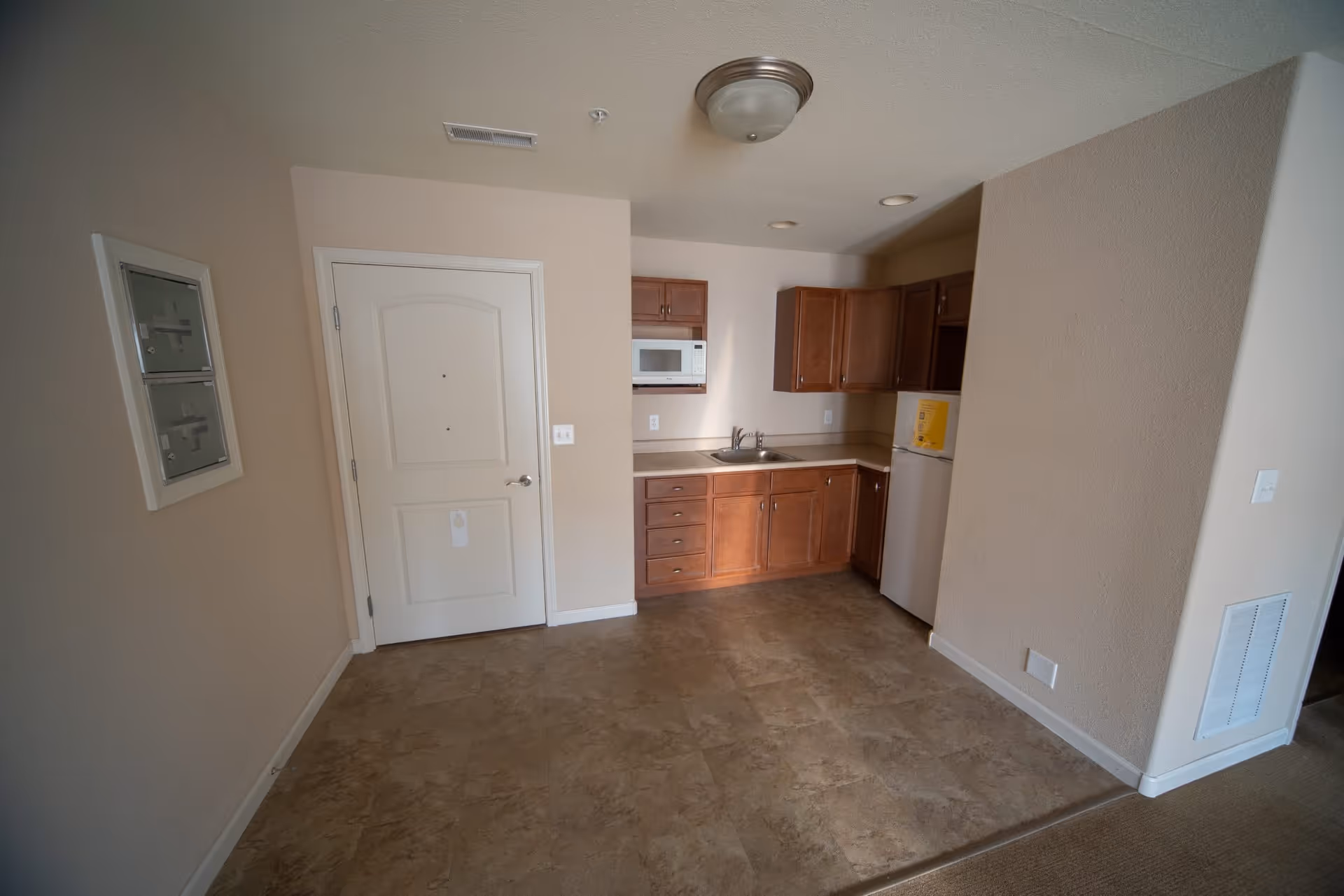 Interior view of a small kitchen area in a senior living facility with wooden cabinets, a microwave, a sink, and a refrigerator. The room has beige walls and tiled flooring, with a white door on the left side and a ceiling light fixture.