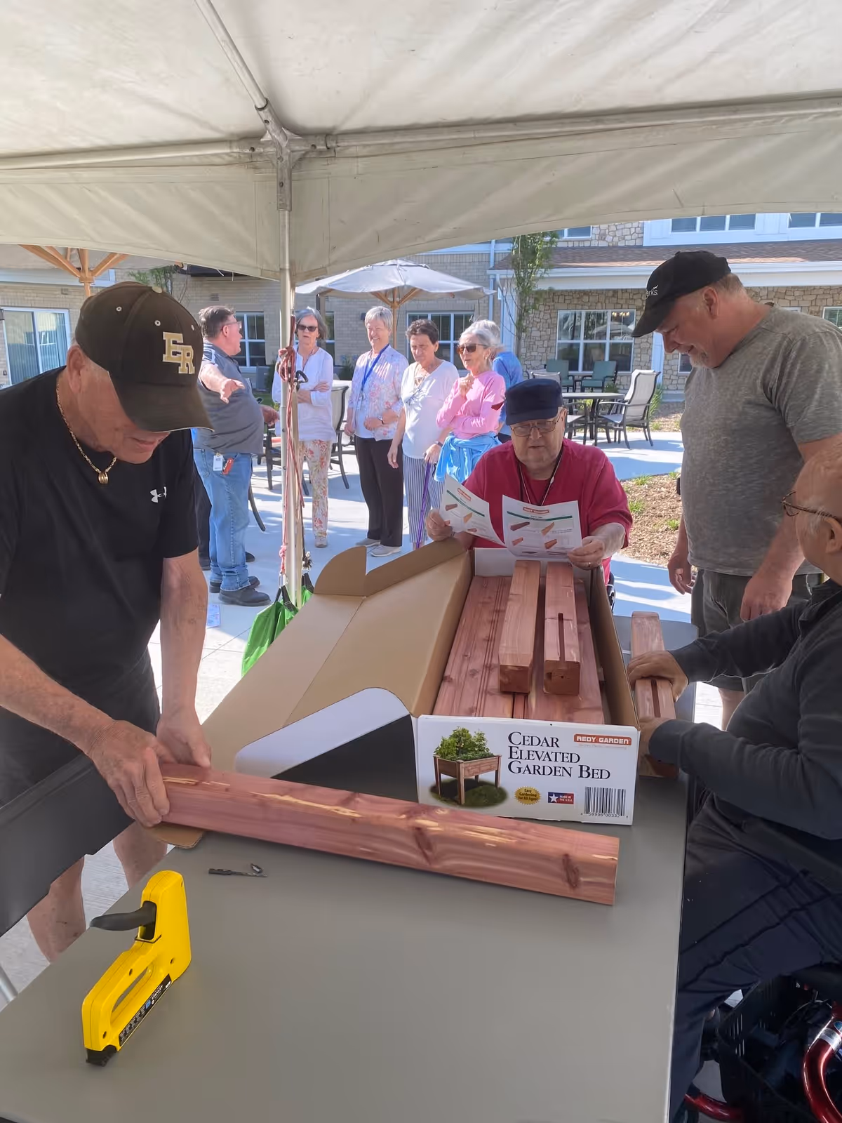 A group of elderly people gathered under a white canopy outdoors, assembling a cedar elevated garden bed. One man is holding a wooden plank, another is reading instructions, and others are standing or sitting around the table with garden bed parts. The background shows a senior living facility building with patio furniture and umbrellas.
