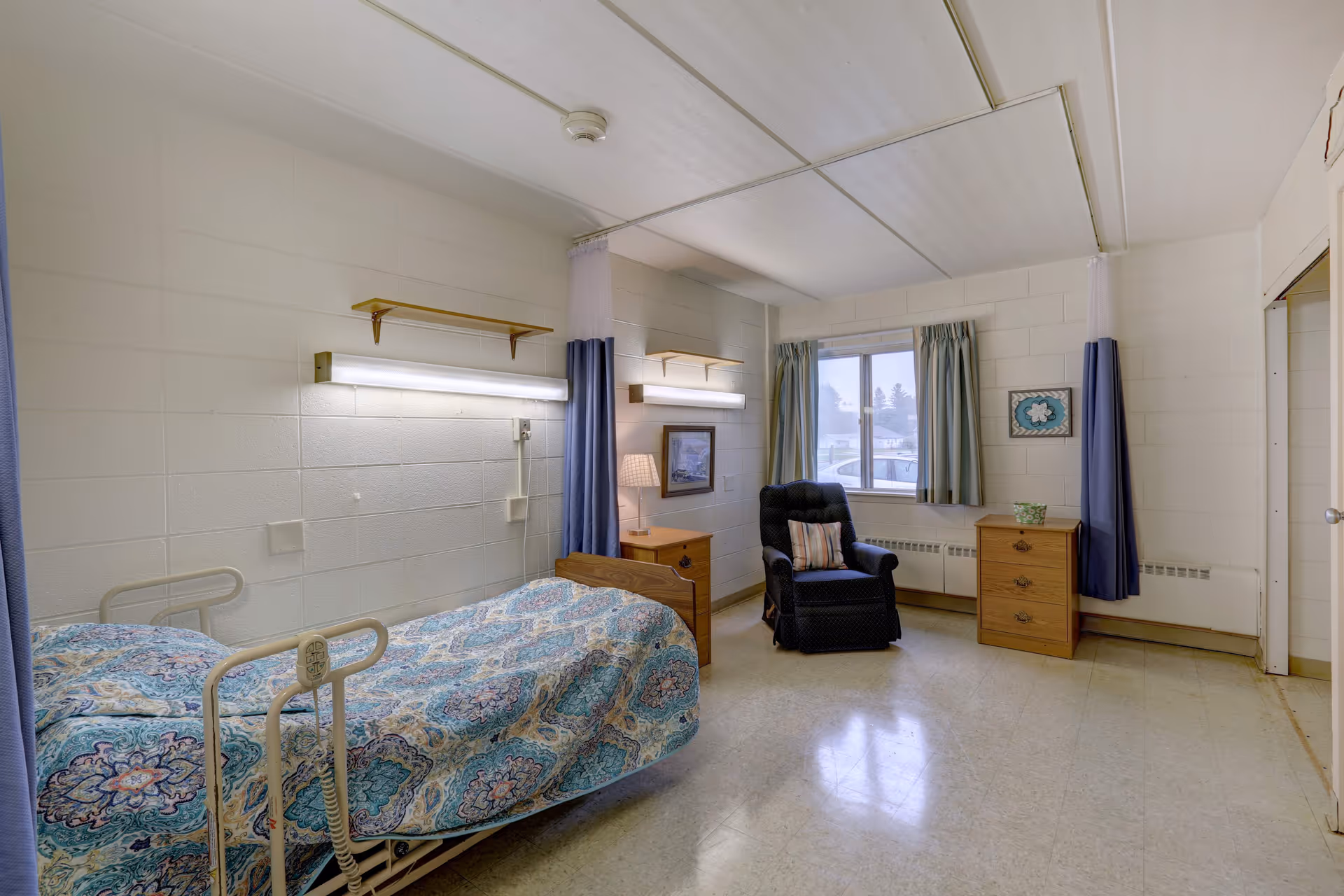 A simple, clean patient room with a single hospital bed covered in a patterned blue and white bedspread. The room has white cinder block walls, two wooden nightstands, a black recliner chair with a striped pillow, a window with green curtains, and two wall-mounted fluorescent lights above the bed. There are privacy curtains on either side of the bed and a framed picture on the wall.