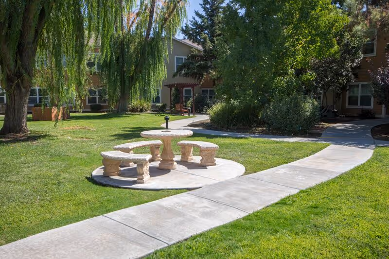Outdoor area with a circular stone table and four matching stone benches on a concrete pad, surrounded by green grass, trees, and shrubs. A paved walkway curves around the seating area leading to a two-story building in the background.