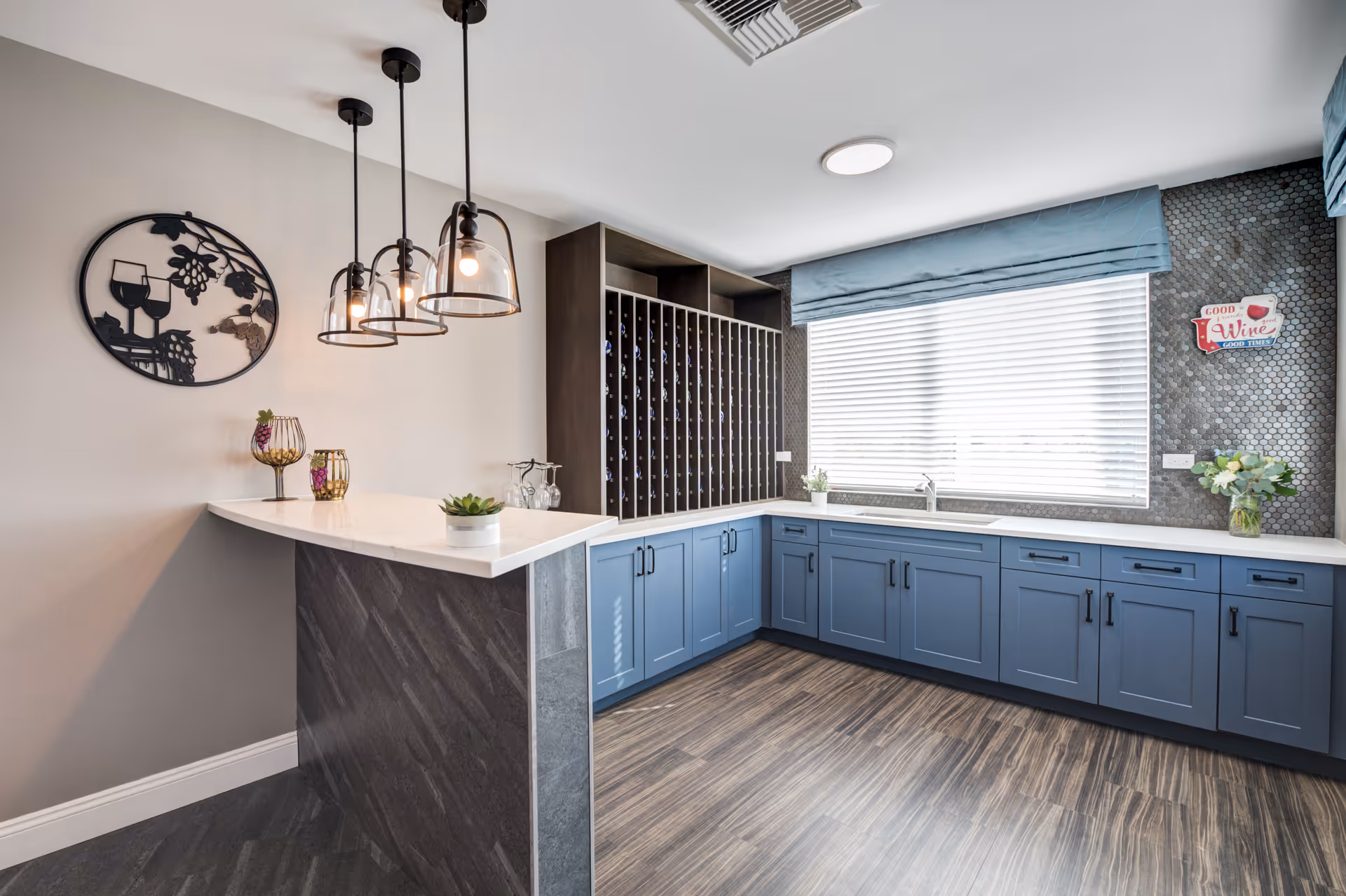 Modern kitchenette with blue cabinetry, a white countertop bar with hanging pendant lights, a wine rack and a large window.