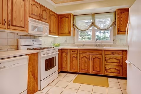 A kitchen with wooden cabinets, white appliances including a stove, microwave, dishwasher, and refrigerator. There is a double window above the sink with sheer curtains, beige tiled backsplash and floor, and a yellow floor mat in front of the sink.