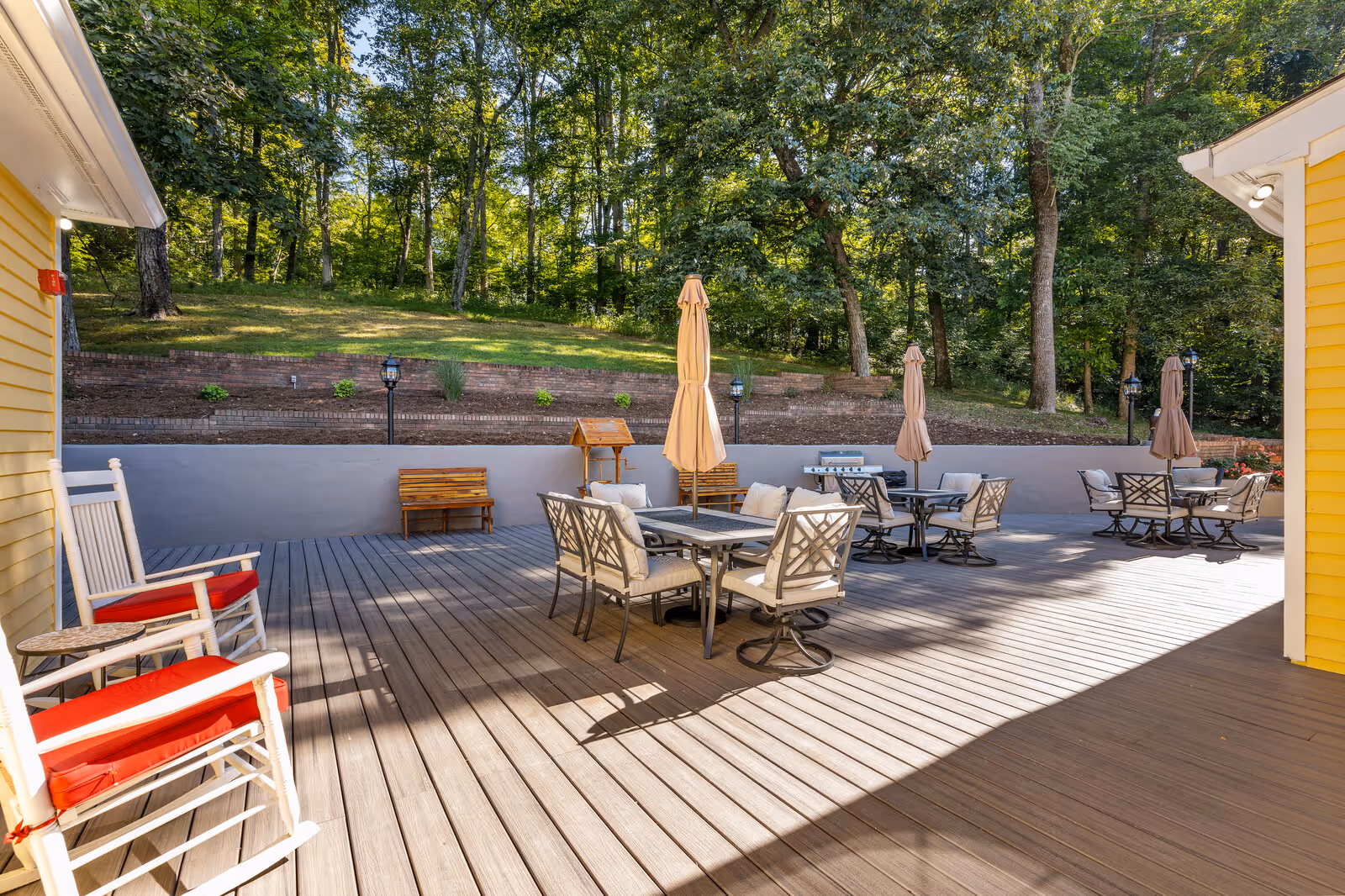 Outdoor patio area with wooden deck featuring several tables with chairs and closed umbrellas. Two white rocking chairs with red cushions are positioned on the left side. The patio is surrounded by a low wall and backed by a grassy slope with trees in the background.