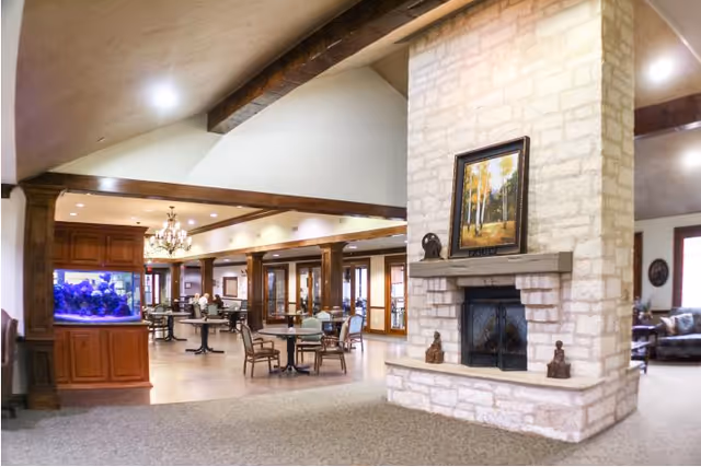 Interior view of a senior living facility common area featuring a stone fireplace with a framed painting above it, several tables and chairs arranged for dining or socializing, wooden beams on the ceiling, a large aquarium on the left, and a chandelier hanging from the ceiling.