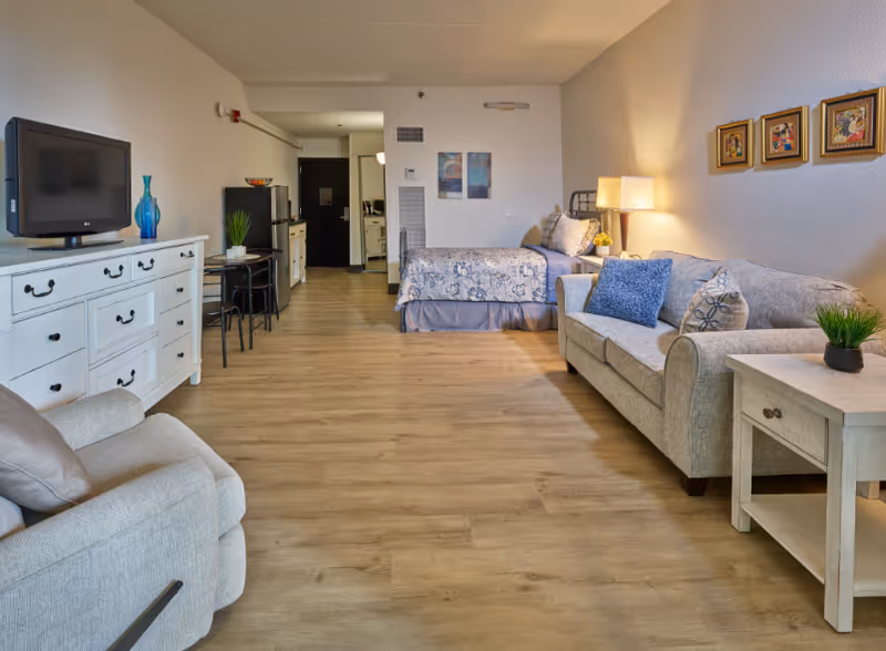 A spacious assisted living studio apartment featuring a bed with floral bedding, a beige sofa with decorative pillows, a matching armchair, a white dresser with a flat-screen TV on top, a small dining table with two chairs, and a kitchenette area in the background. The room has wooden flooring and light-colored walls adorned with framed artwork and a table lamp.