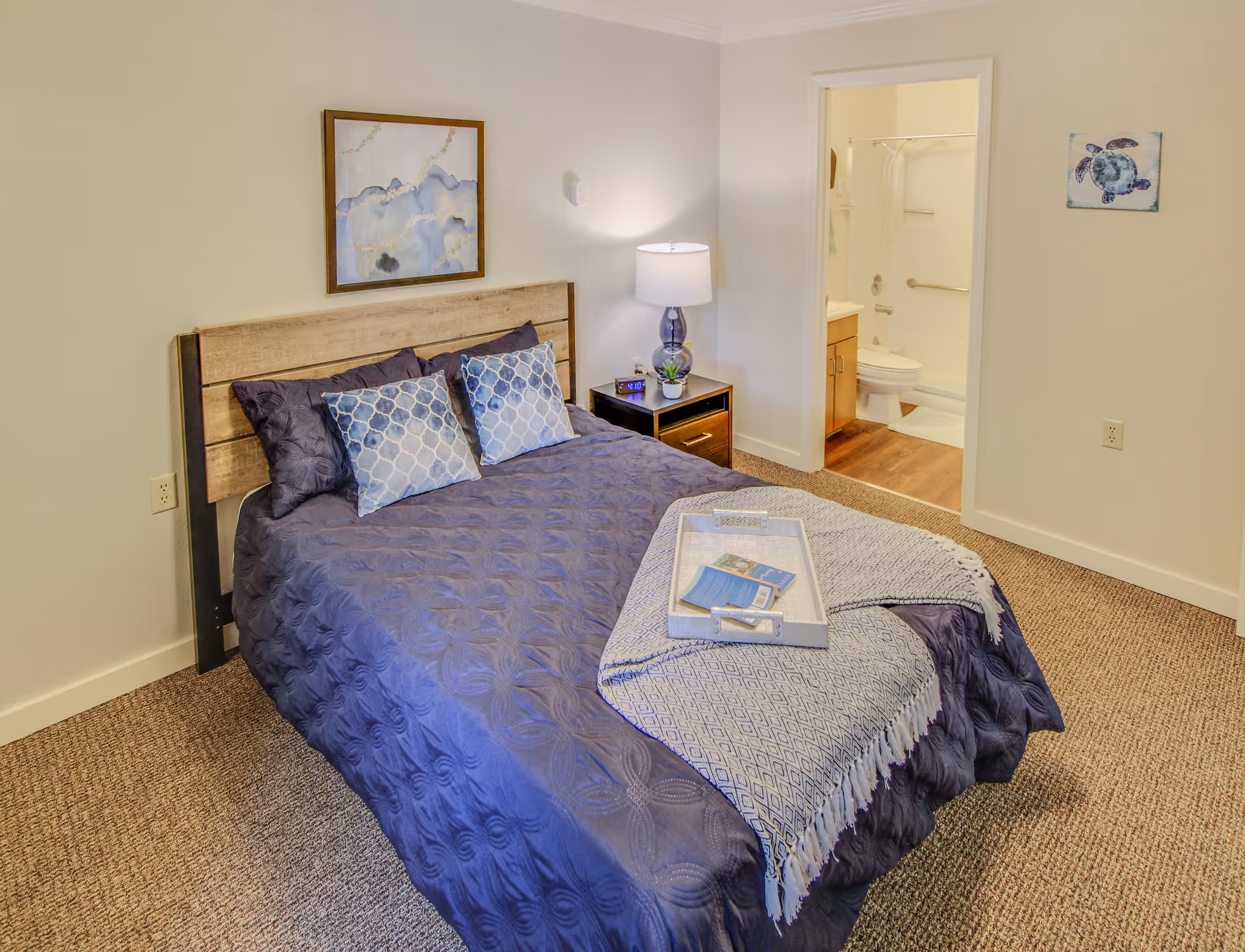 A bedroom with a bed covered in a dark blue quilt and two patterned pillows. A small nightstand with a lamp and a small plant is next to the bed. There is a framed abstract painting on the wall above the bed. An open doorway reveals a bathroom with a toilet, sink, and bathtub with a shower curtain. The room has beige carpet and light-colored walls.