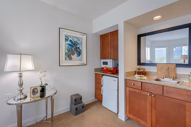 Small kitchenette and seating area with wooden cabinets, granite countertop and sink, microwave and mini fridge, plus a side table with a lamp and artwork on the wall.
