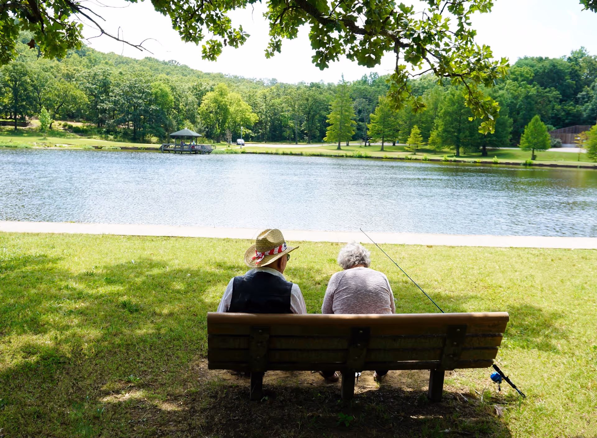 Two elderly people sitting on a wooden bench by a lake, with one person wearing a straw hat and the other holding a fishing rod. The scene is surrounded by green grass, trees, and a small gazebo across the water under a clear sky.