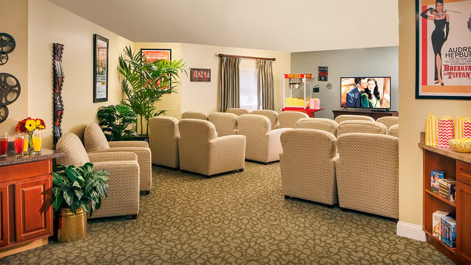 A cozy movie theater room with multiple beige armchairs arranged in rows facing a television screen showing a movie scene. There is a popcorn machine and popcorn containers on a counter to the right. The room is decorated with movie-themed posters, plants, and a small refreshment area with drinks and flowers on the left side.