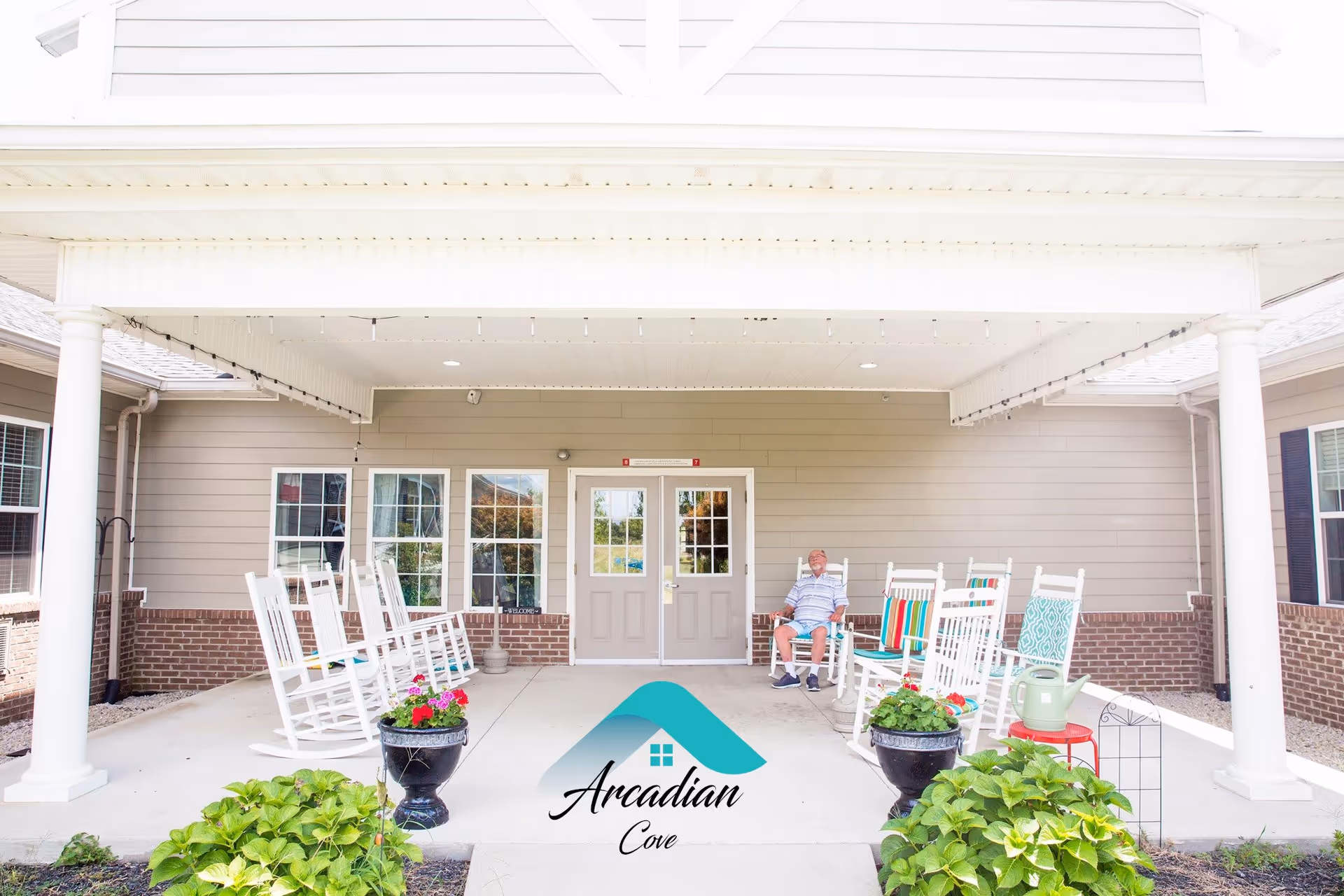 Covered porch area at Arcadian Cove Senior Living with white rocking chairs arranged on both sides. A man is seated on one of the rocking chairs on the right side. There are potted plants with flowers in front of the porch and a watering can on a small red table. The building exterior features beige siding with brick accents and white columns supporting the porch roof.
