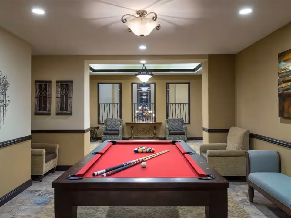 A cozy interior lounge featuring a red-felt pool table centered with armchairs and additional seating around it.