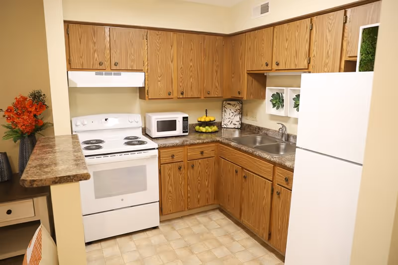 A compact kitchen with wooden cabinets, a white electric stove with oven, a white microwave, a double sink, and a white refrigerator. The countertop has a two-tiered fruit stand with lemons and green apples, a decorative cutting board, and two small wall-mounted shelves with green plants. There is a small counter extension on the left side with a vase of orange flowers and a small table with a drawer below it.