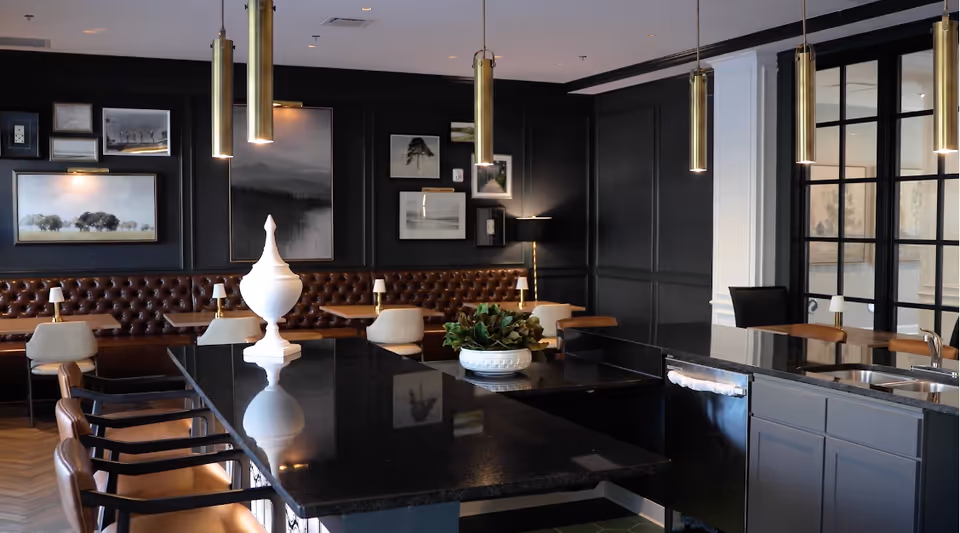 A modern dining area with a large black granite island countertop surrounded by brown leather chairs. The background features a dark paneled wall adorned with various framed artworks and a brown tufted leather bench with small tables and beige chairs. Pendant lights hang from the ceiling, and there is a sink and dishwasher integrated into the island.