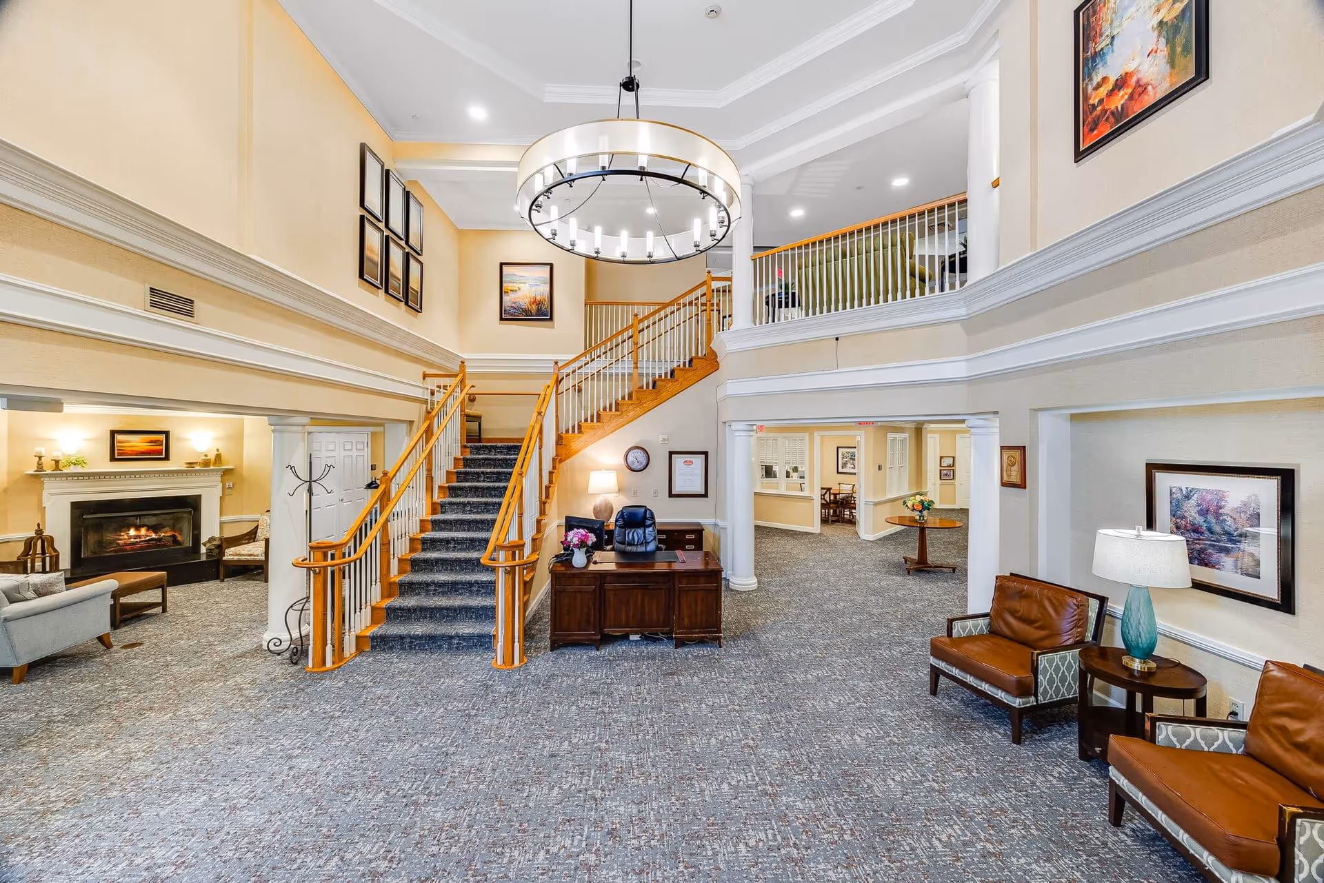 Spacious and well-lit senior living facility lobby with a central staircase leading to an upper floor. The area features a large circular chandelier, comfortable seating including leather chairs and sofas, a fireplace, framed artwork on the walls, and a reception desk with a chair and lamp. The carpeted floor and warm color tones create a welcoming atmosphere.