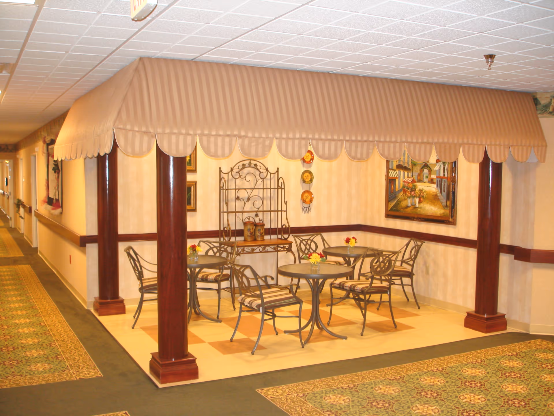 Indoor seating area in a senior living facility with a canopy supported by four wooden columns. The area contains three round tables with metal chairs that have striped cushions. The floor beneath the seating area is tiled, and the surrounding floor is carpeted with a patterned design. The walls are decorated with framed artwork and decorative plates.