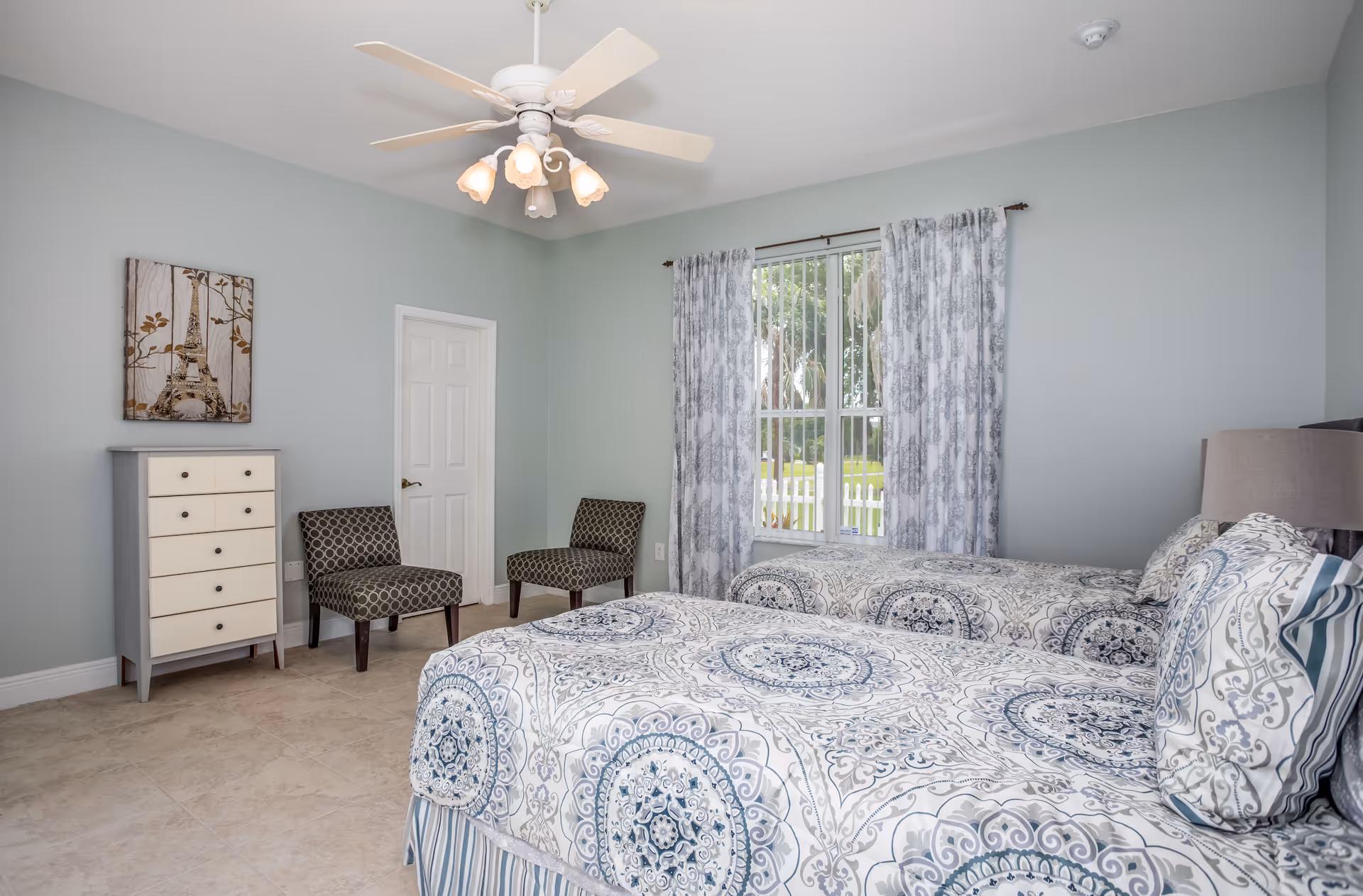 A bright bedroom with two twin beds featuring patterned blue and white bedding. The room has light blue walls, a ceiling fan with lights, a window with patterned curtains, two patterned chairs, a white door, and a small dresser with a picture of the Eiffel Tower above it.
