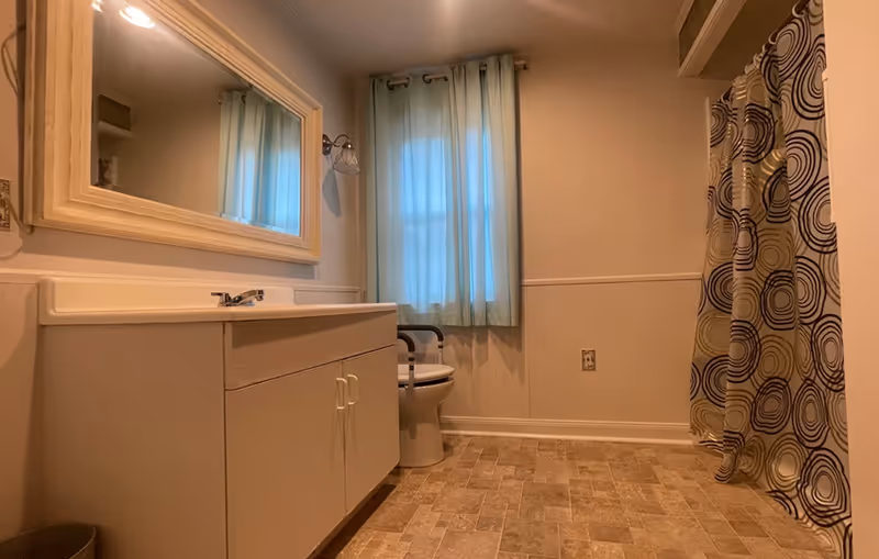 Spacious bathroom with a vanity and mirror on the left, a toilet beneath a curtained window in the center, and a shower with a patterned curtain on the right.