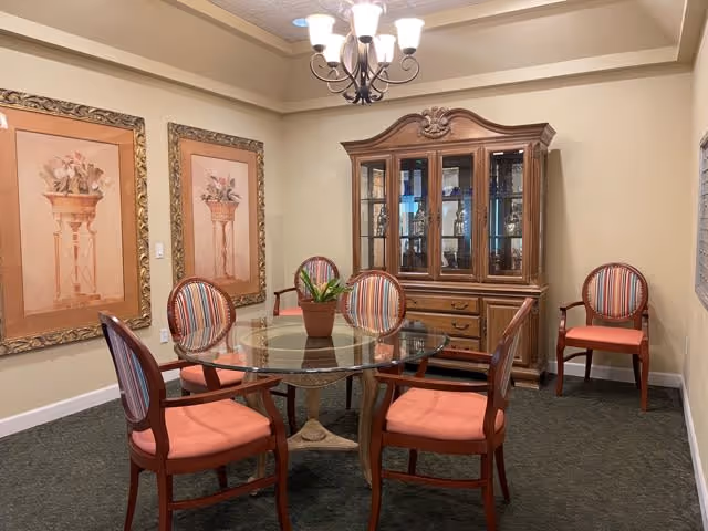 A dining room with a round glass table surrounded by five wooden chairs with coral-colored cushions and striped backs. A wooden china cabinet with glass doors is against the wall, displaying glassware. Two framed floral paintings hang on the left wall, and a chandelier with five lights hangs from the ceiling. The room has beige walls and a dark carpeted floor.
