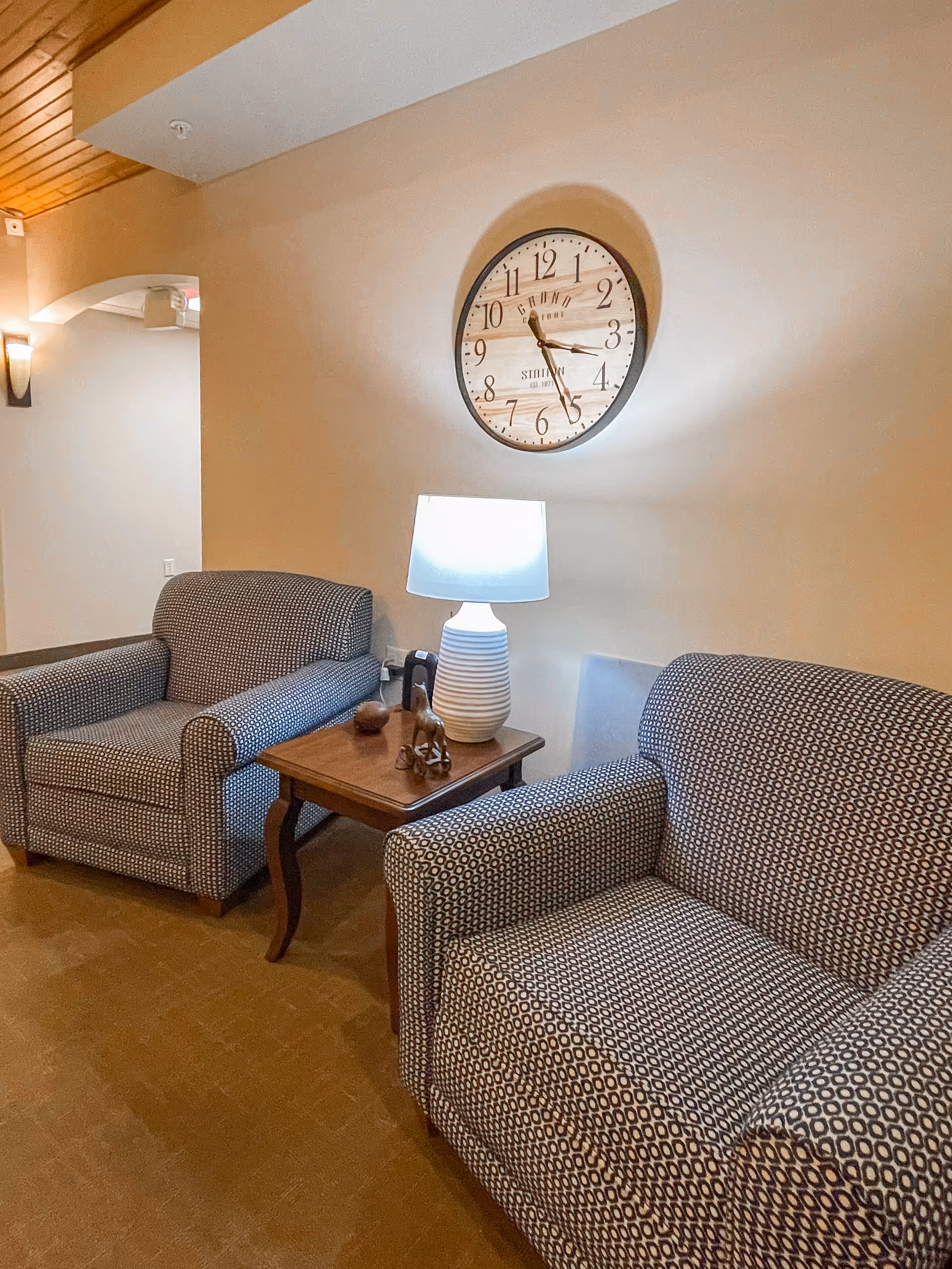 Two patterned armchairs flank a wooden side table with a lamp beneath a large wall clock in a cozy sitting area.