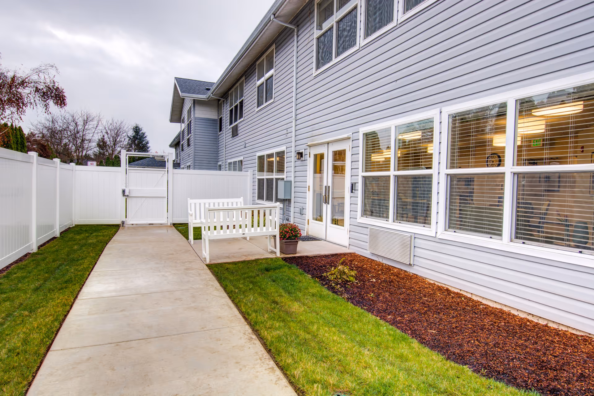 Paved walkway beside a gray assisted living building with large windows, a white fence, and a bench in a small courtyard.
