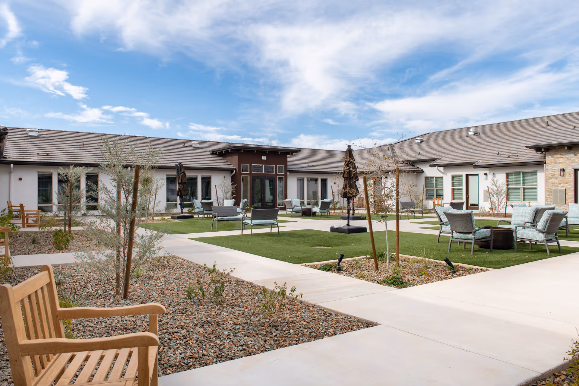 Sunny landscaped courtyard with walkways, seating areas, and a single-story senior living building in the background.