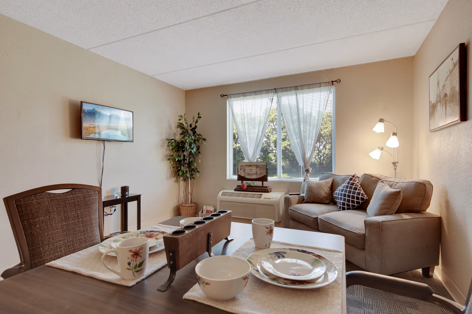 A cozy living room area with a small dining table set for two, featuring floral-patterned dishes and mugs. The room has a beige couch with several pillows, a floor lamp with three lights, a potted plant in the corner, a wall-mounted TV, and a window with sheer white curtains letting in natural light.