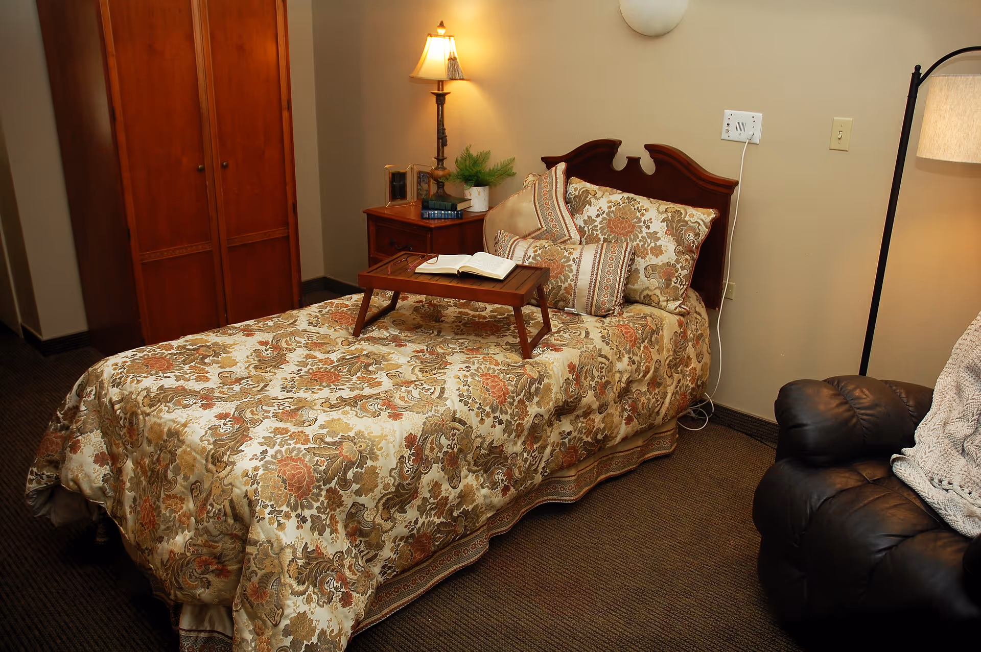A cozy bedroom with a single bed covered in a floral patterned bedspread and matching pillows. A wooden tray with an open book rests on the bed. Next to the bed is a wooden nightstand with a lamp, a small plant, and some books. There is a wooden wardrobe in the corner and a dark leather recliner chair with a knitted blanket on it. The walls are beige and the floor is carpeted.