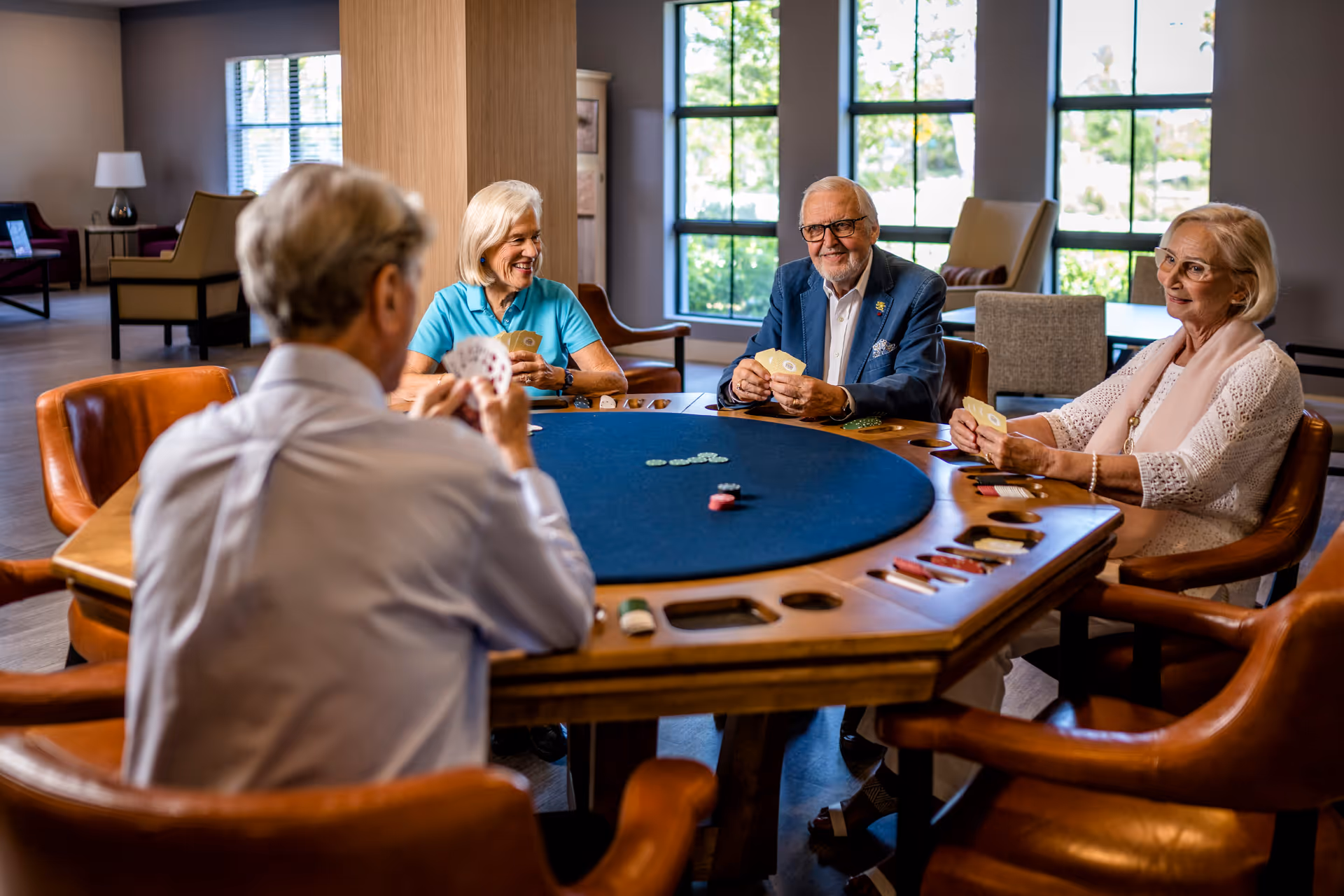 Four elderly people sitting around a poker table in a well-lit room with large windows, playing cards and smiling. The room has comfortable chairs and a relaxed atmosphere.