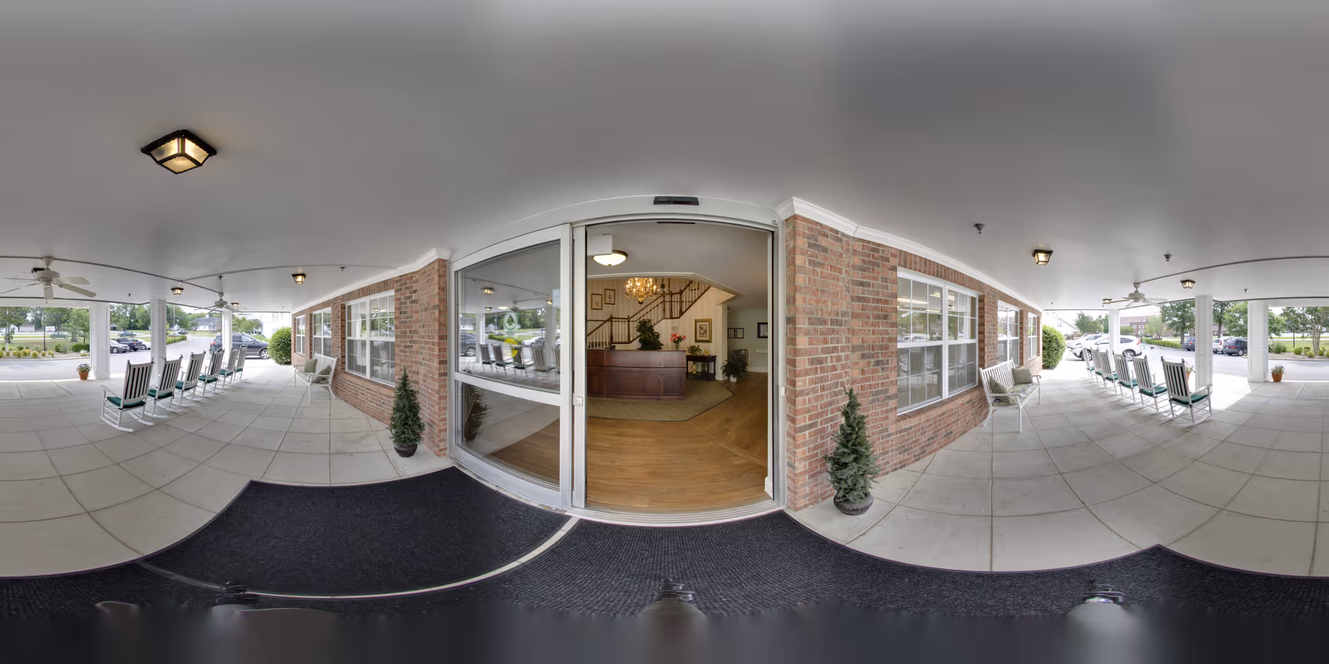 Covered front entrance and porch of a senior living facility with rocking chairs, brick facade, and glass doors opening to a lobby.