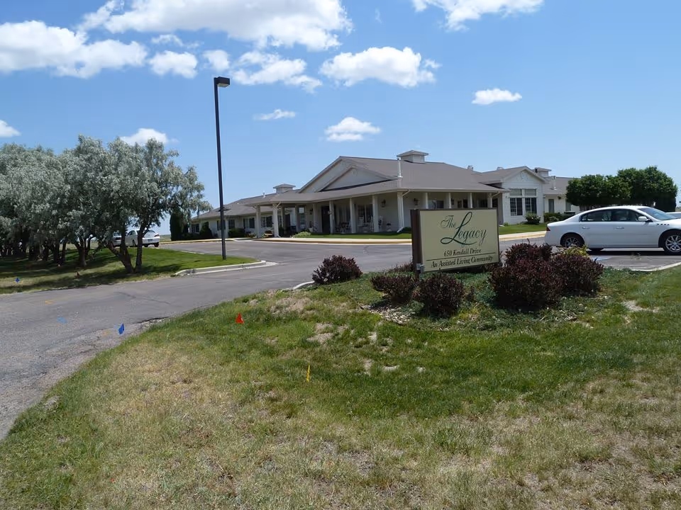 Front exterior of The Legacy senior living building with an entrance canopy, sign, parked car, and landscaped lawn under a blue sky.
