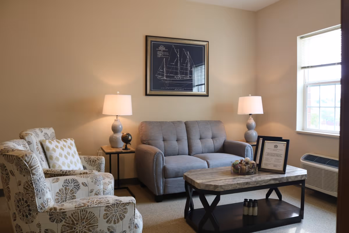 A cozy living room with a gray loveseat, a patterned armchair with a decorative pillow, two matching table lamps on side tables, a framed blueprint of a sailboat on the wall, a window with blinds, and a coffee table with decorative items and framed certificates.