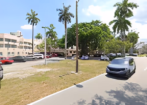 Outdoor view of a parking area with several cars parked on a grassy and paved surface. There are tall trees and a multi-story building in the background under a partly cloudy sky.