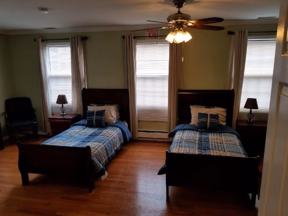 A bedroom with two single beds, each with blue and white plaid bedding and pillows. Between the beds are two wooden nightstands, each with a lamp. There are three windows with blinds and beige curtains behind the beds. A ceiling fan with lights is mounted on the ceiling. A dark chair is placed in the corner of the room.