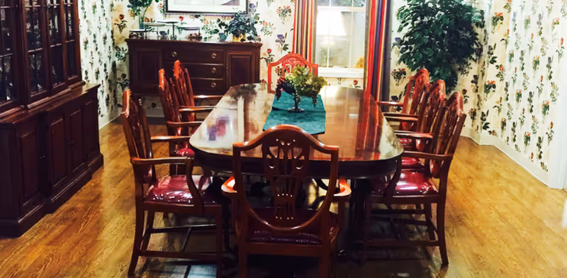 A traditional dining room with a long polished wooden table surrounded by eight wooden chairs with red cushioned seats. The room has floral wallpaper, a wooden sideboard with decorative items, a large window with curtains, and a wooden floor.
