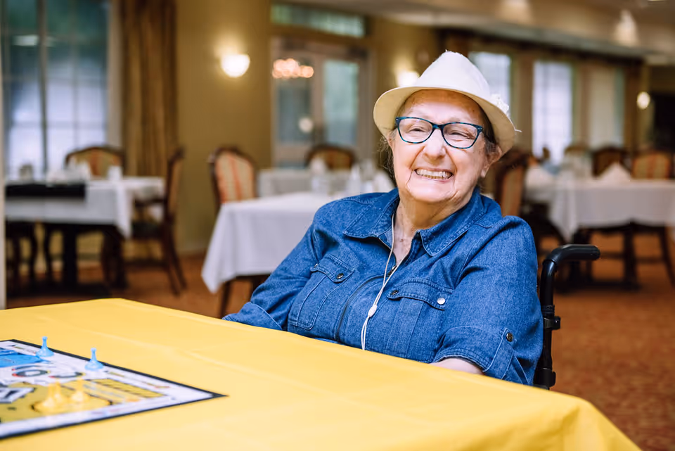An elderly woman wearing a white hat, glasses, and a denim shirt is smiling while sitting in a wheelchair at a table covered with a yellow tablecloth. A board game is partially visible on the table. The background shows a dining area with tables and chairs, softly lit with wall sconces and windows.