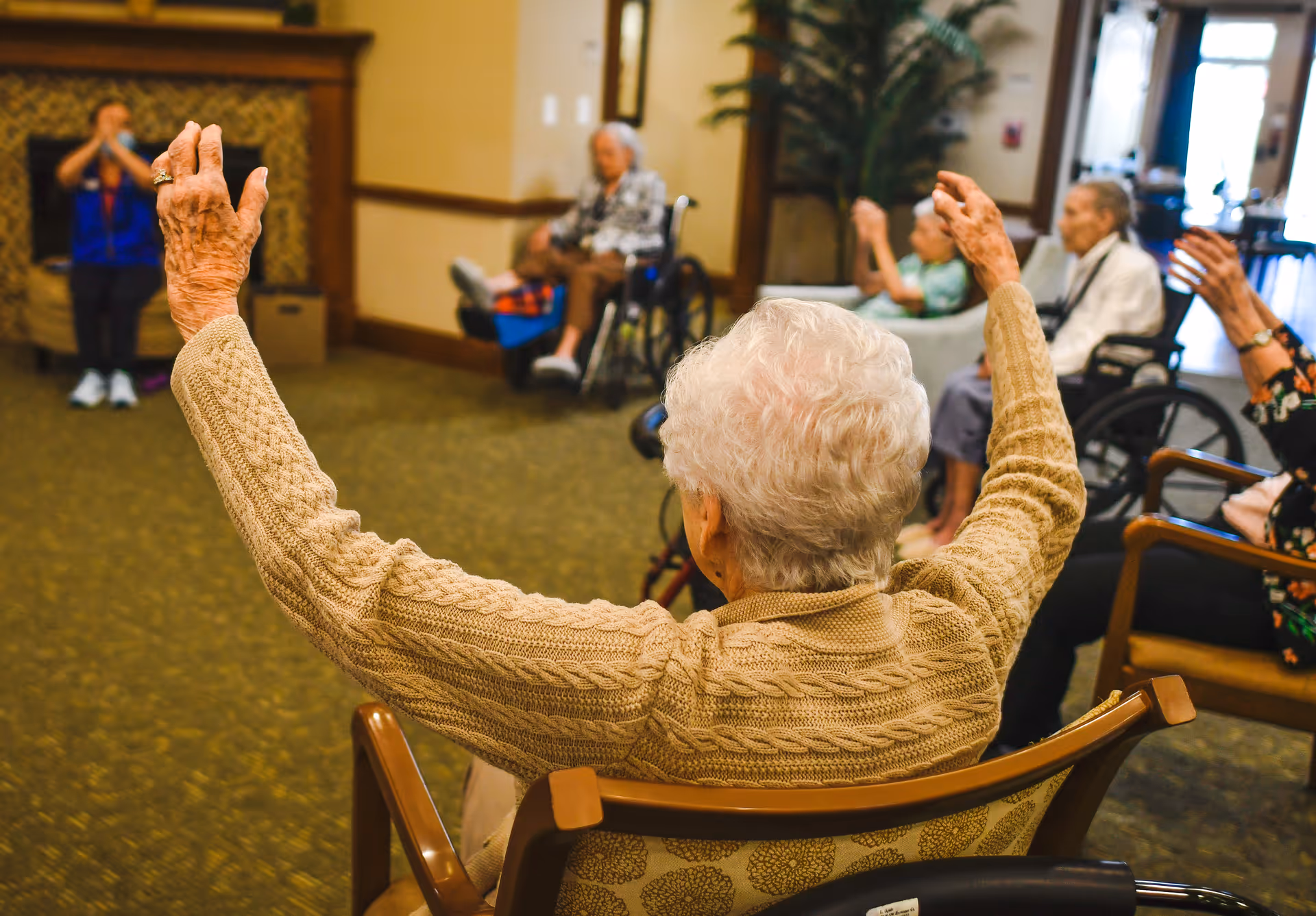 A group of elderly individuals sitting in chairs and wheelchairs in a common room, participating in a seated exercise or stretching activity led by an instructor in the background.
