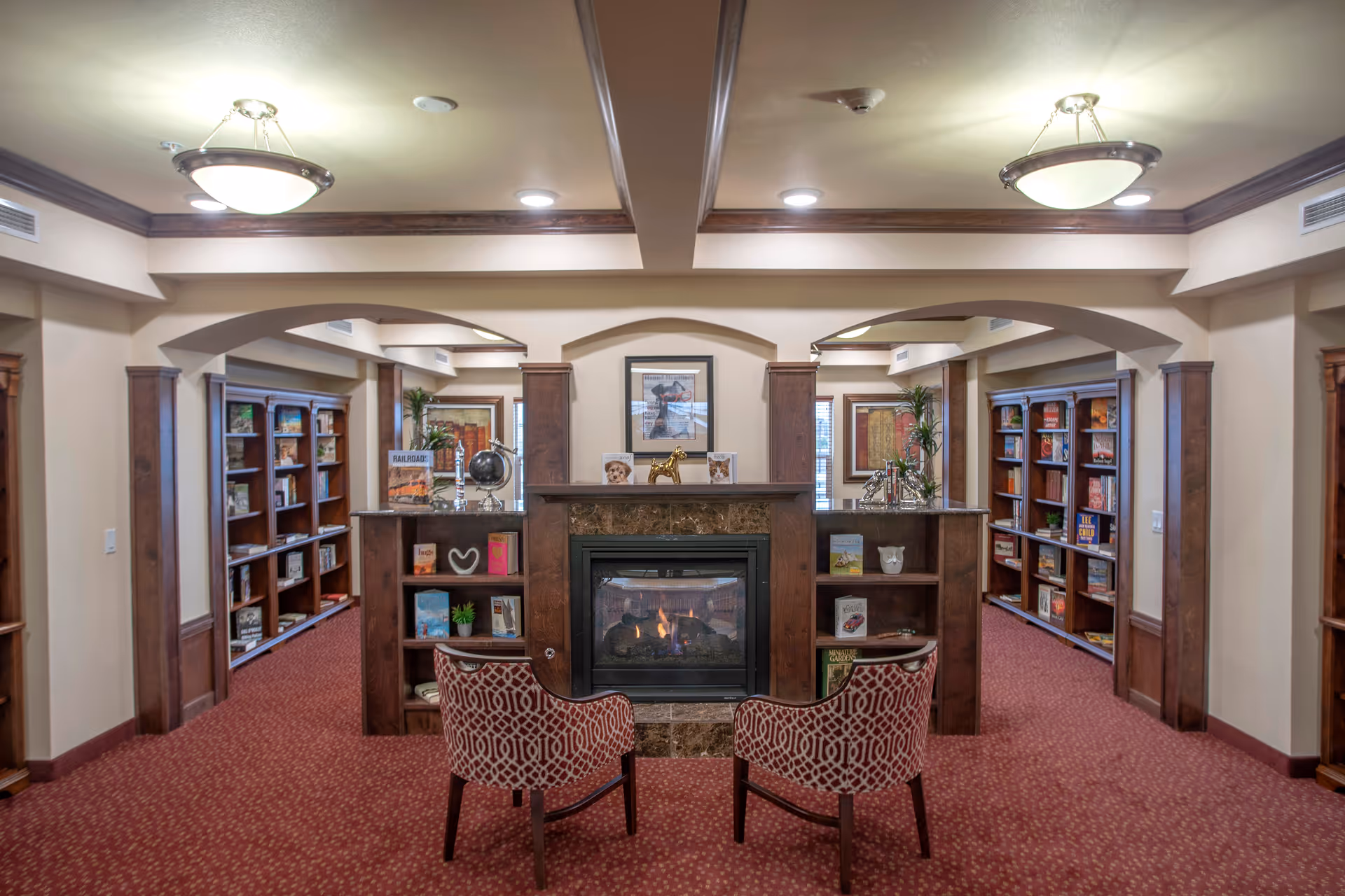 A cozy library or reading room with a central fireplace surrounded by wooden shelves filled with books and decorative items. Two patterned armchairs face the fireplace, and the room features warm lighting with ceiling fixtures and recessed lights. The carpet is red with a subtle pattern, and there are framed pictures and plants on the walls and shelves.