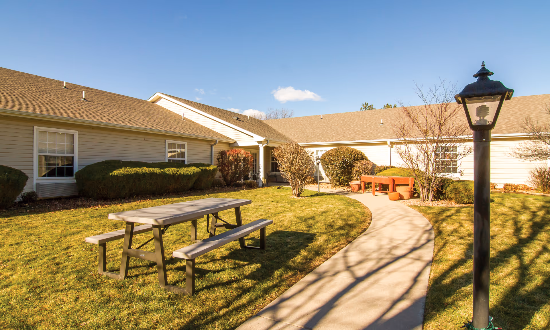 Sunny courtyard with a picnic table, benches, paved walkway, lamp post, and single-story building surrounding the lawn.