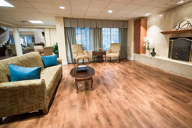 A spacious living room with wooden flooring, a patterned sofa with blue cushions, two armchairs with a small round table between them, large windows with curtains, and a fireplace with decorative items on the mantel.