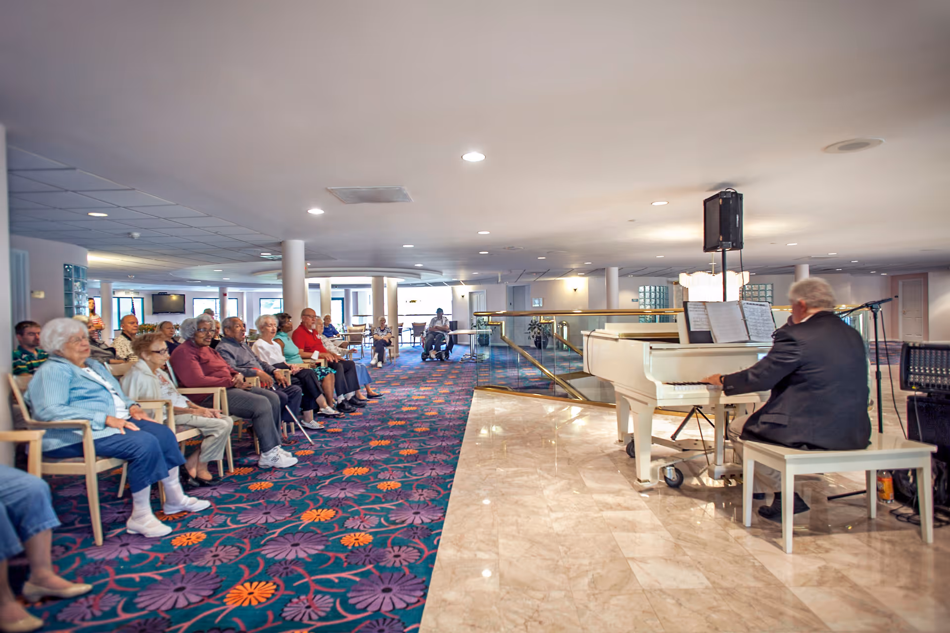 A group of elderly people seated in chairs along a colorful carpeted area, attentively watching a man playing a white grand piano in a spacious, well-lit room with marble flooring and white pillars.
