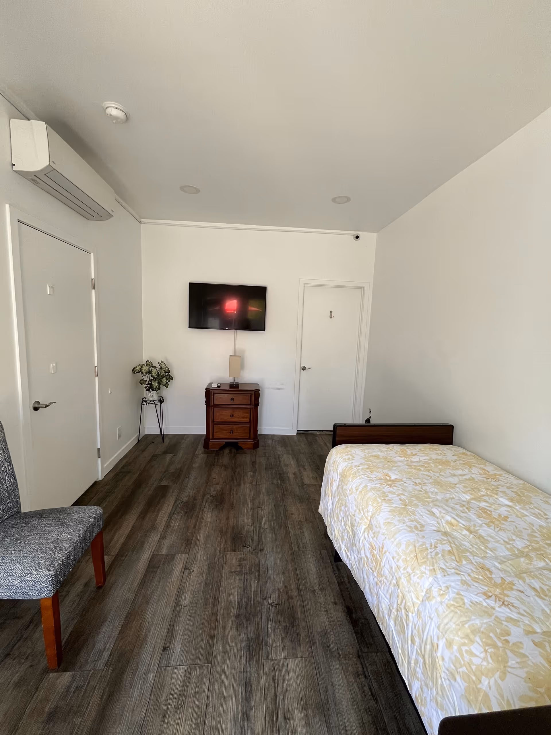 A simple bedroom with a single bed covered in a yellow and white floral bedspread, a wooden nightstand with a lamp, a flat-screen TV mounted on the wall, a gray patterned chair, and a small potted plant on a stand. The room has wood flooring, white walls, and two closed white doors.