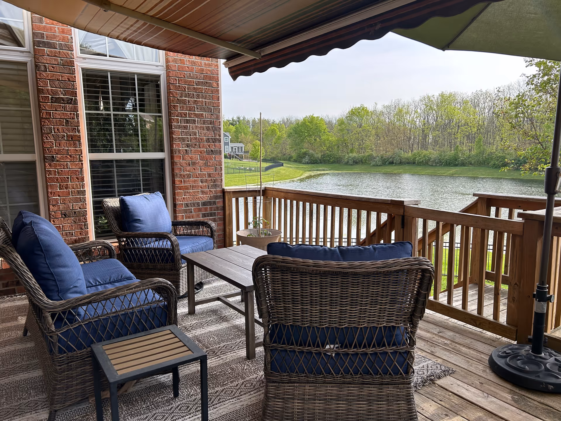 A wooden deck patio with wicker furniture including three chairs with blue cushions and a small table. The deck overlooks a pond with green trees in the background. There is a large umbrella providing shade over the seating area.