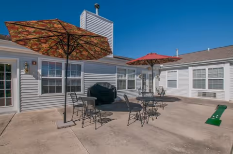 Sunlit outdoor courtyard patio with metal tables and chairs, colorful umbrellas, a grill, and surrounding building windows.
