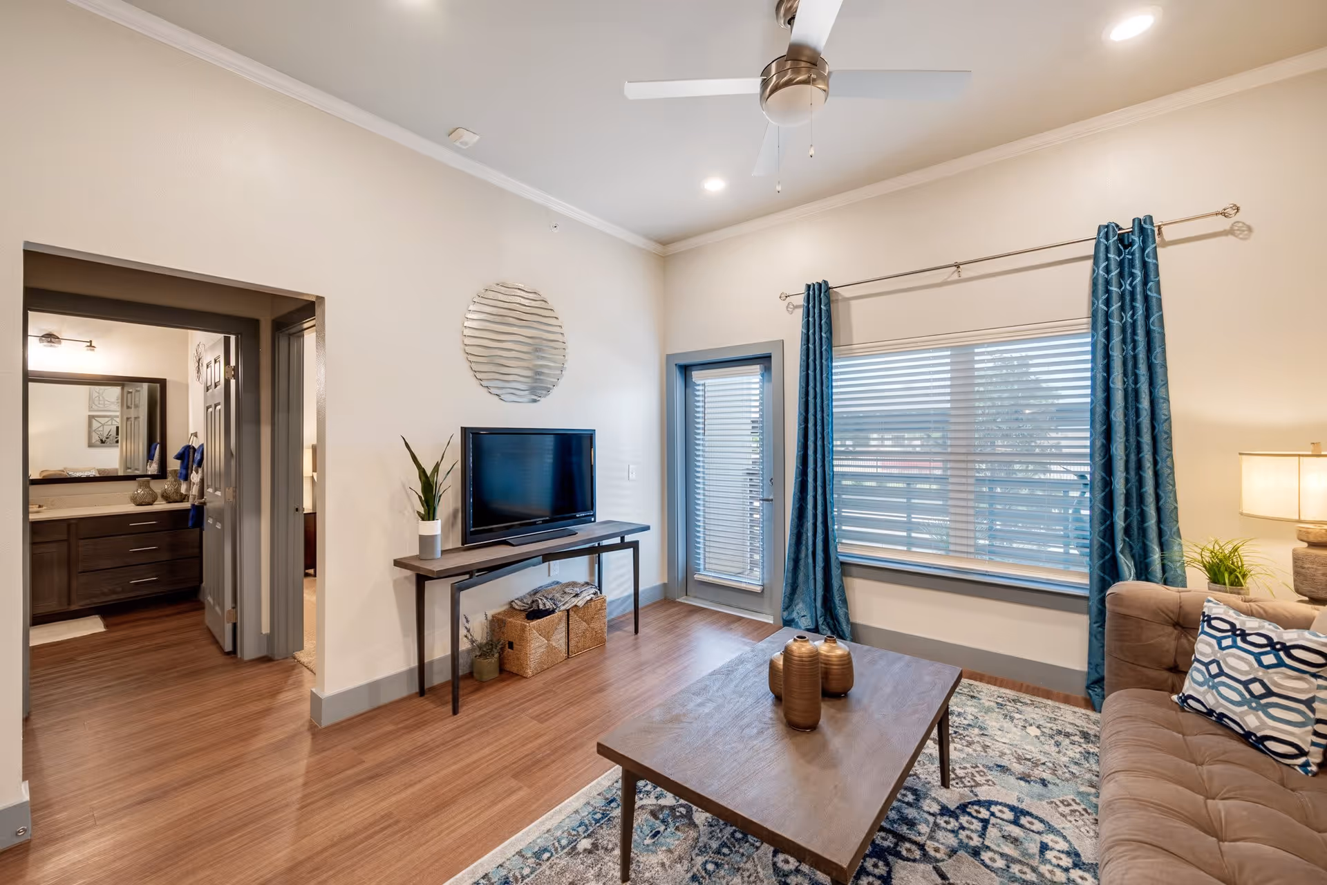 A cozy living room in a senior citizen home featuring a brown sofa with patterned cushions, a wooden coffee table with decorative vases, a flat-screen TV on a slim console table, and a large window with blue curtains. The room has wooden flooring, a ceiling fan, and an open doorway leading to a bathroom and bedroom.