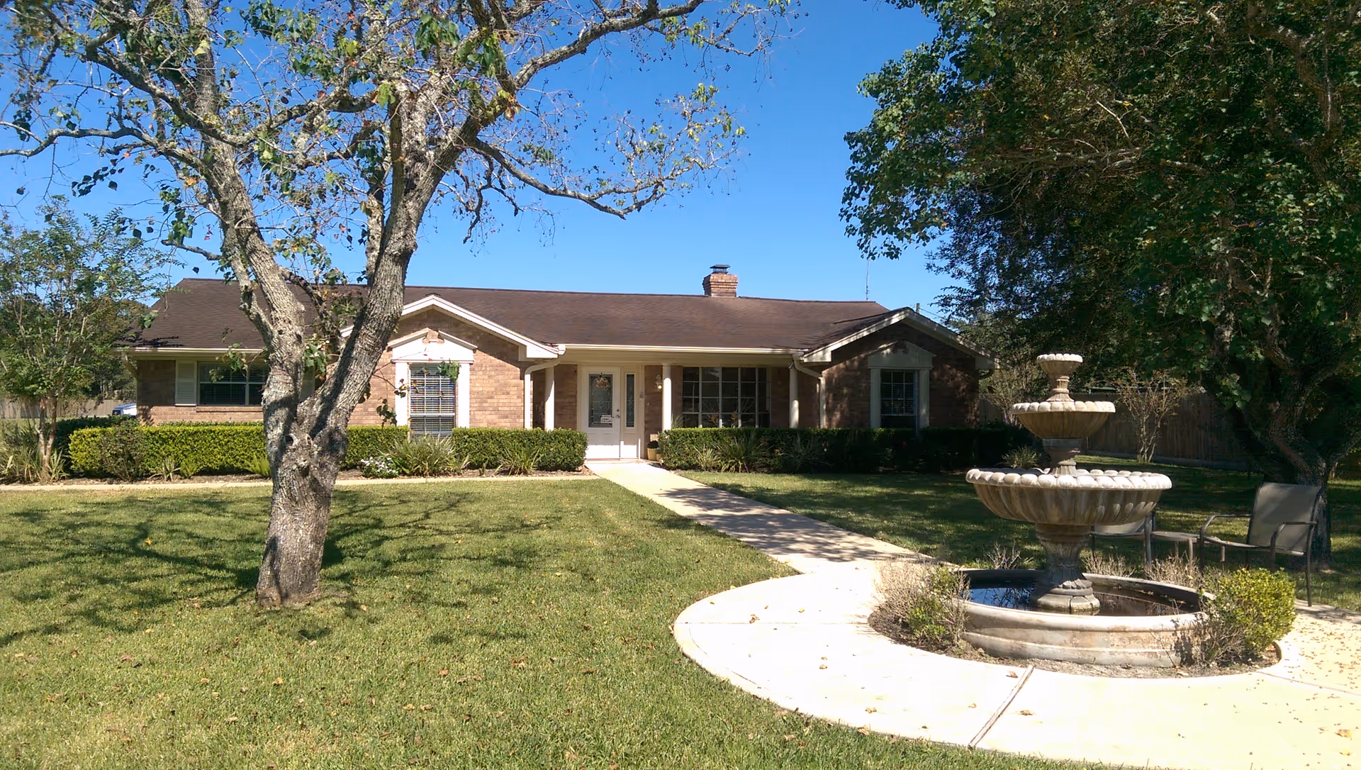 Single-story brick building with a front lawn, curved walkway, trees and a tiered fountain.