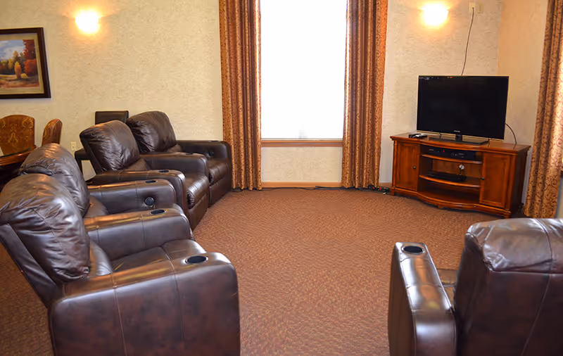 A cozy living room with four dark brown leather recliners arranged facing a flat-screen TV on a wooden TV stand. The room has beige walls, a window with patterned curtains, and a framed painting on the wall. The carpet is brown with a subtle pattern.