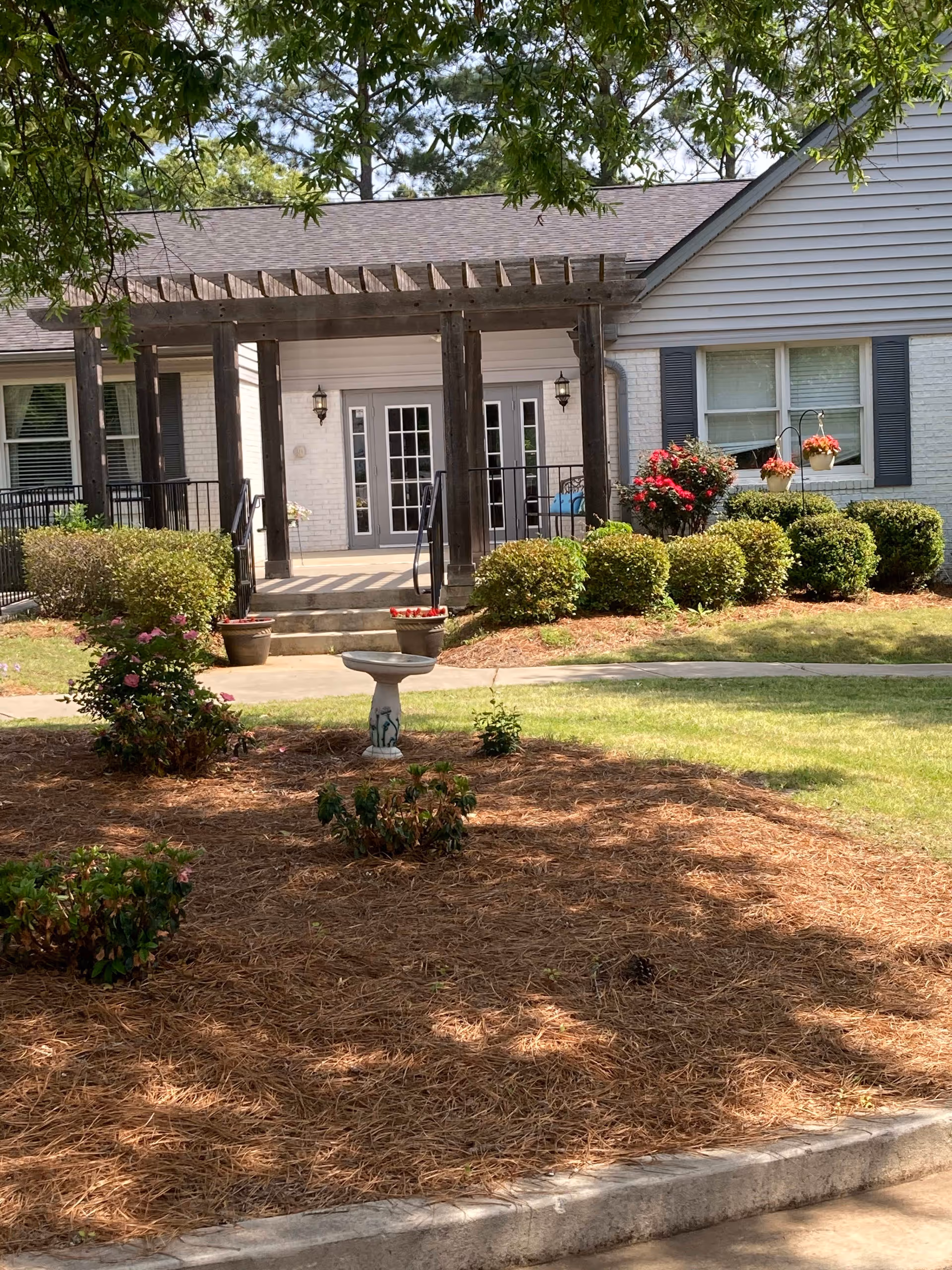 Outdoor view of a senior living facility with a garden area featuring bushes, flowers, and a birdbath in the foreground. The building has a white exterior with a covered porch supported by wooden beams and double glass doors. Trees provide shade over the garden.