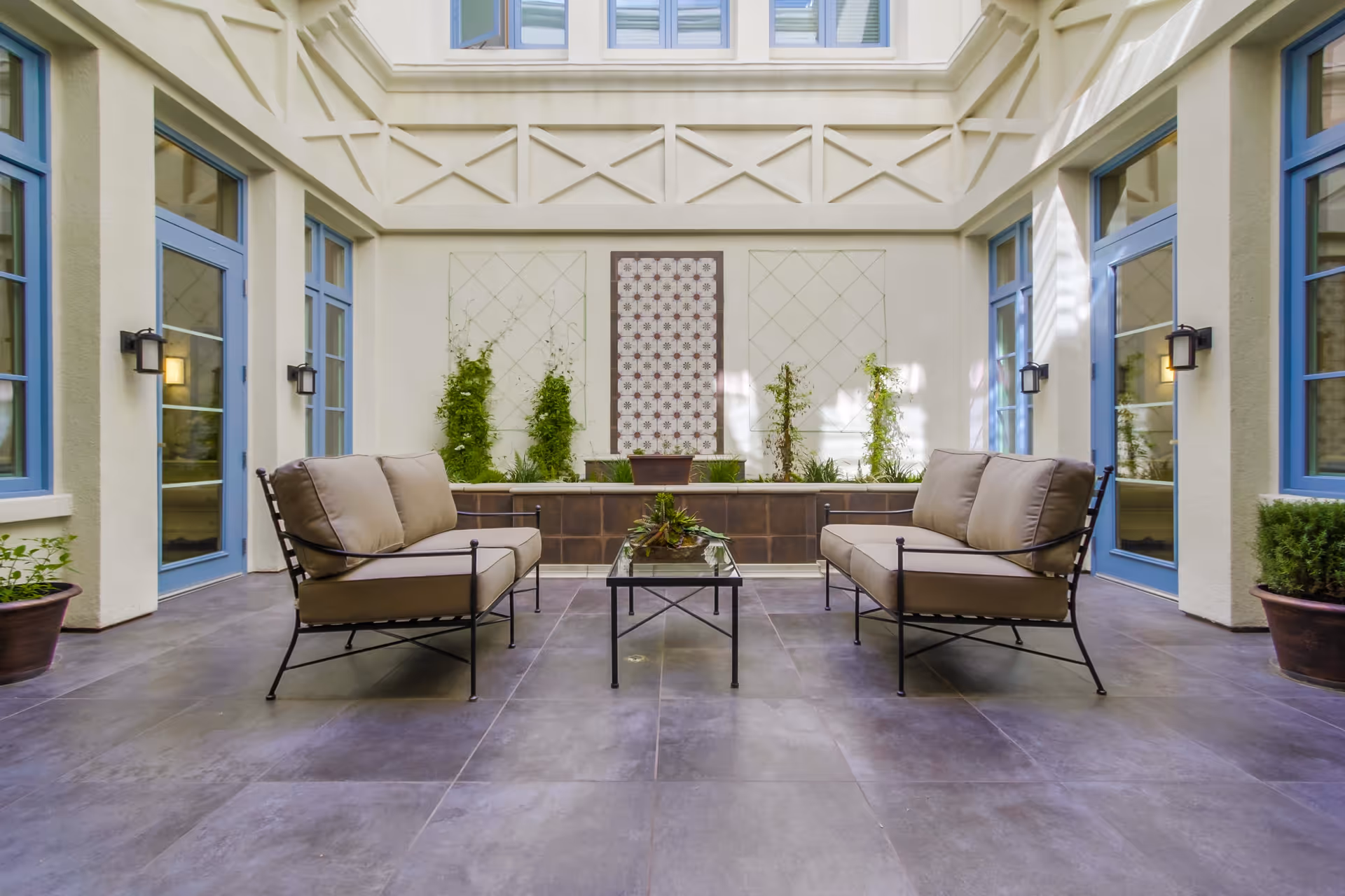Indoor seating area with two cushioned metal sofas facing each other and a glass coffee table in between. The space has large blue-framed windows and doors, potted plants, and a decorative wall with climbing plants and a patterned panel. The floor is tiled in a gray tone.