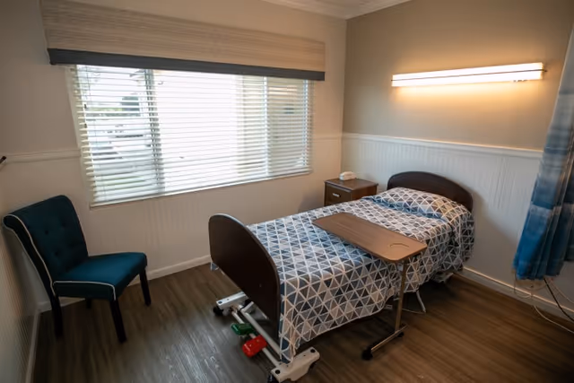 A private patient bedroom with an adjustable single bed, overbed table, nightstand, chair, and a window with blinds.