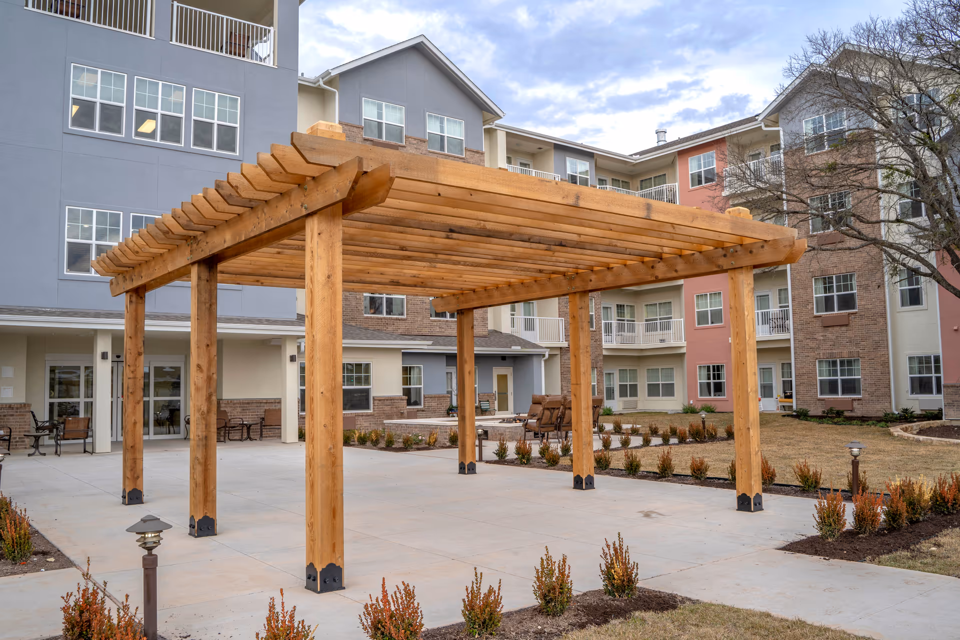 Outdoor courtyard area of a senior living facility featuring a large wooden pergola on a concrete patio surrounded by small landscaped bushes. The background shows a multi-story building with balconies and windows under a partly cloudy sky.